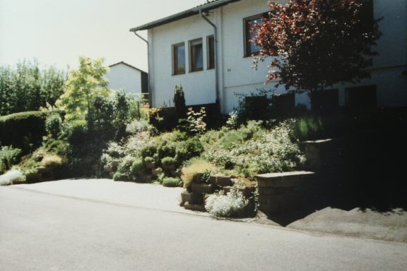 Blühender Garten und Hausfassade mit einer Einfahrt.
