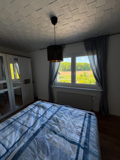 Schlafzimmer mit Bett, Fenster und Vorhängen, Blick ins Grüne.