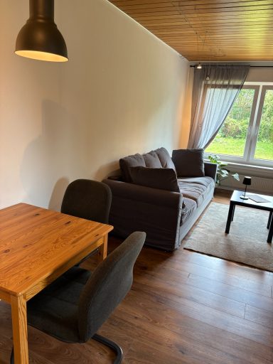 Heller Wohnraum mit Sofa, Tisch und Stühlen, Fenster mit Gardine und Blick ins Grüne.